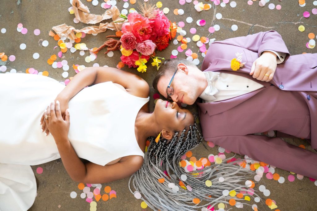 Newlywed couple laying on ground posing with confetti and pink bouquet