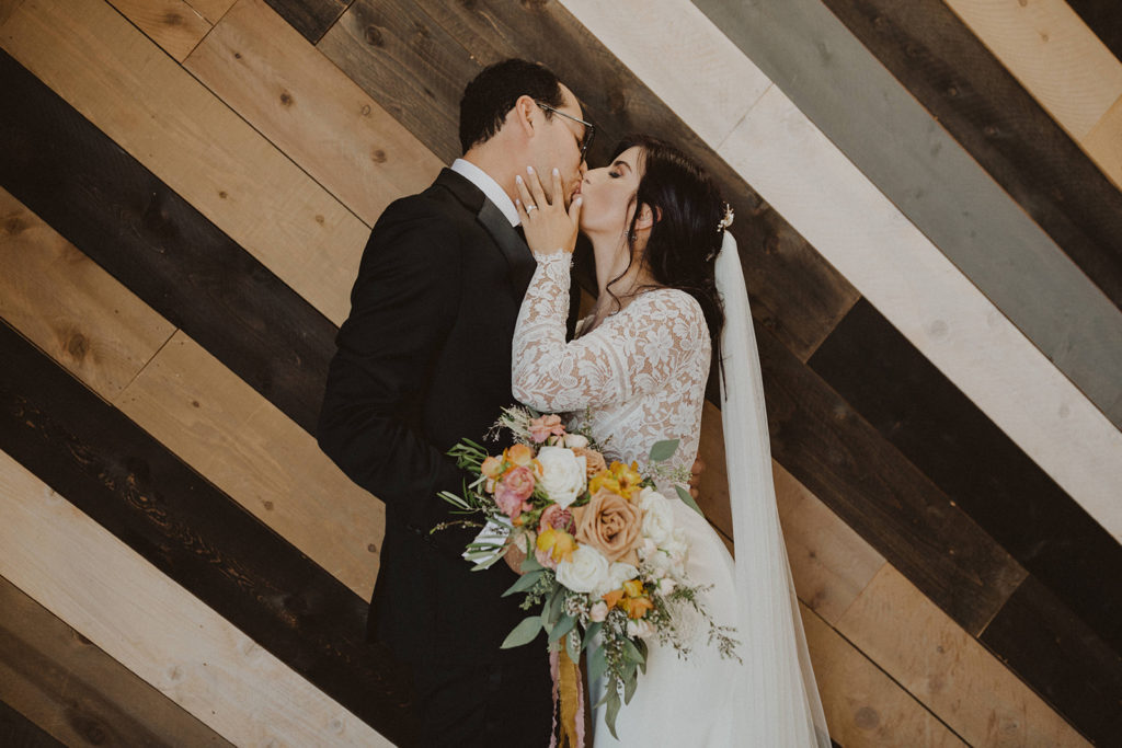 Fall wedding couple with colorful Fall bouquet kissing in the reception area of The Wood Shed in Vista California