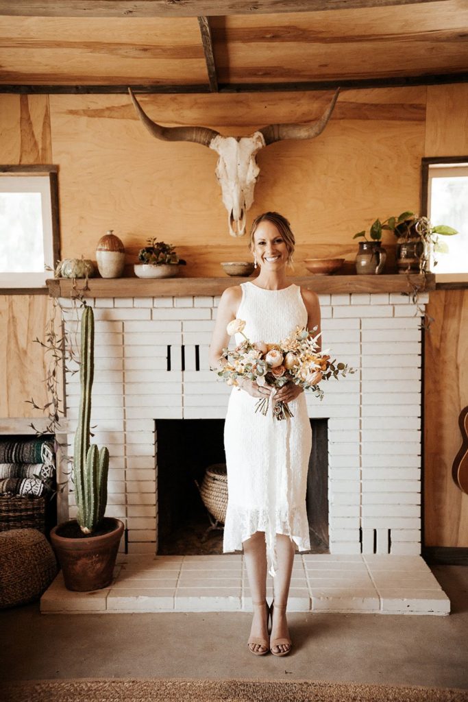 The Caravan house in Joshua Tree bride standing in front of the fireplace with peach and white bridal bouquet