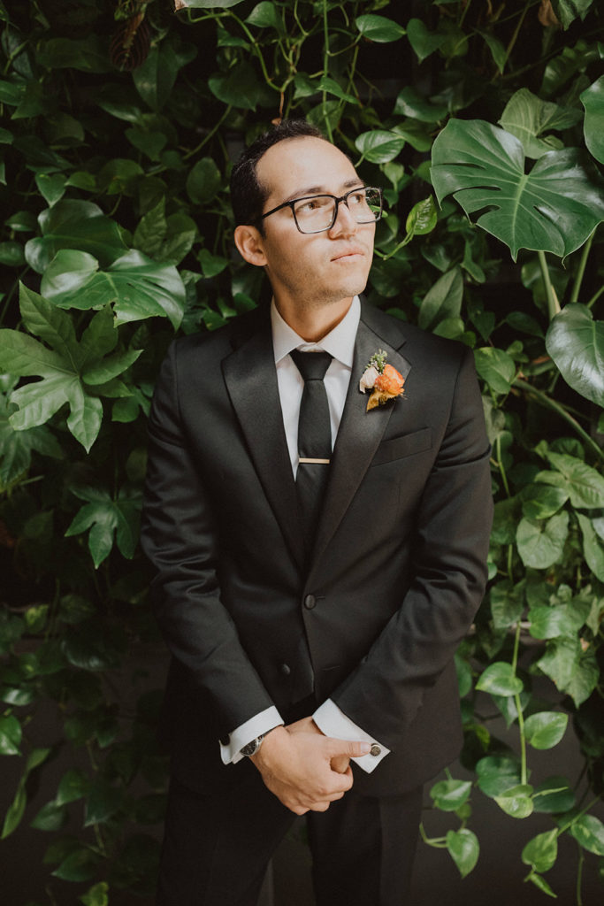 Fall wedding groom in a black suit with an orange boutonniere standing in front of a tropical greenery living wall at The Wood Shed in Vista California