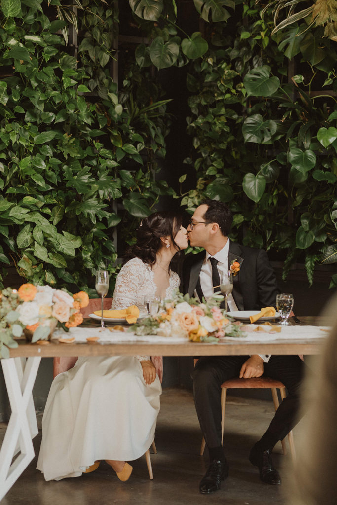 Fall wedding couple kissing at their sweetheart table with colorful Fall flowers and a tropical back drop at The Wood Shed in Vista California