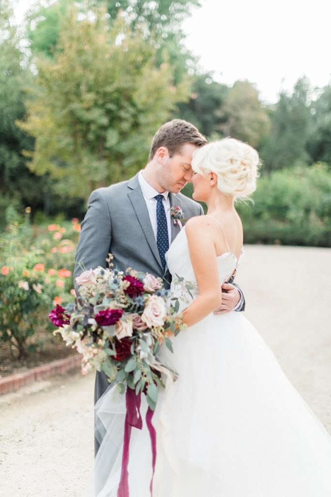 Organic blue and burgundy winter wedding groom in a gray suit and bride holding a large bouquet at Orcutt Ranch