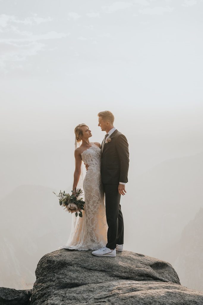Yosemite elopement bride wearing a white lace dress holding a protea bouquet with groom standing on a large rock