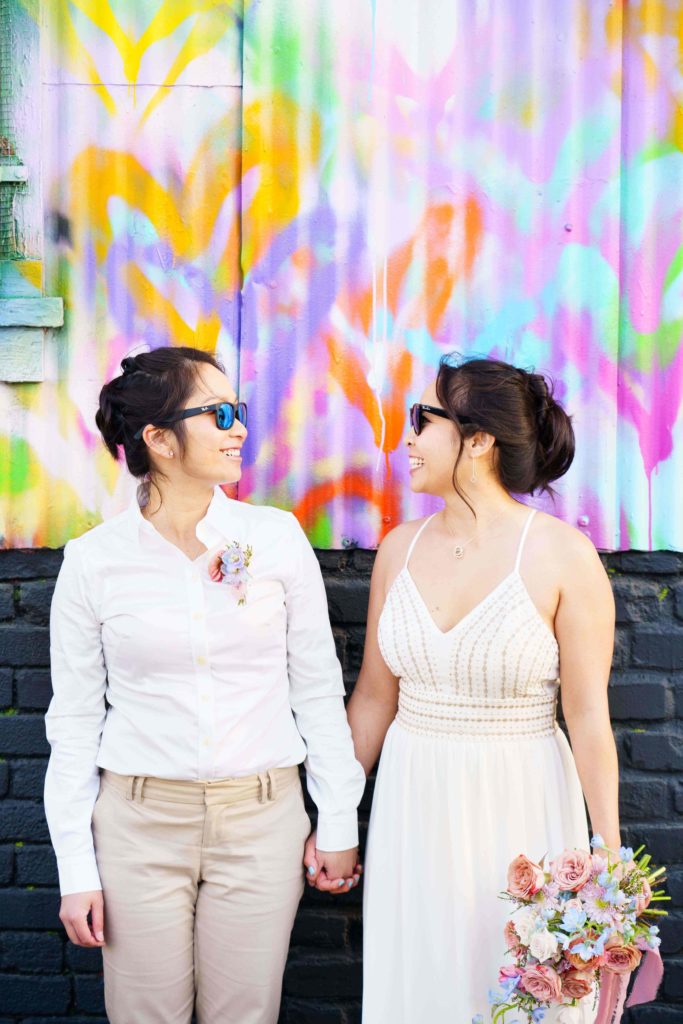 Lesbian couple holding hands in front of a graffiti wall with a colorful bouquet