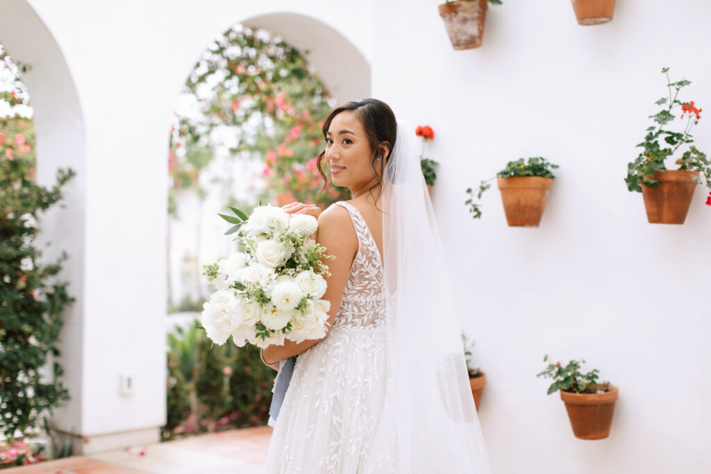 Bride in front of a hanging plant wall holding a large bouquet with white peonies and greenery with blue ribbon at La Ventura Event Center in San Clemente California