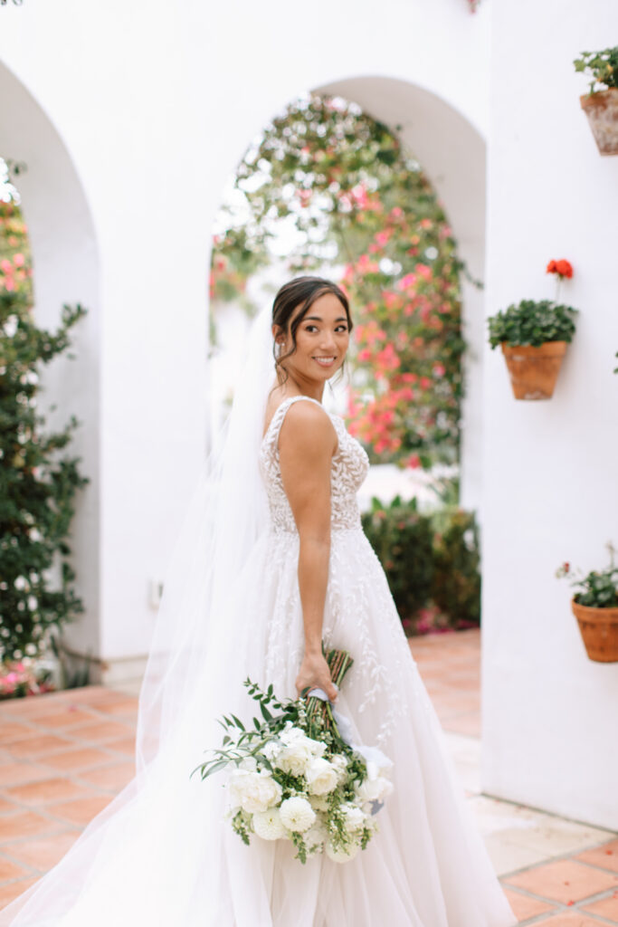 Bride looking behind her and holding a large bouquet with white flowers and greenery at La Ventura Event Center in San Clemente