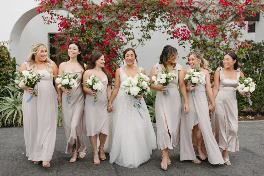 Bride smiling walking with her bridesmaids wearing champagne colored dresses holding bouquets with white flowers at La Ventura Event Center