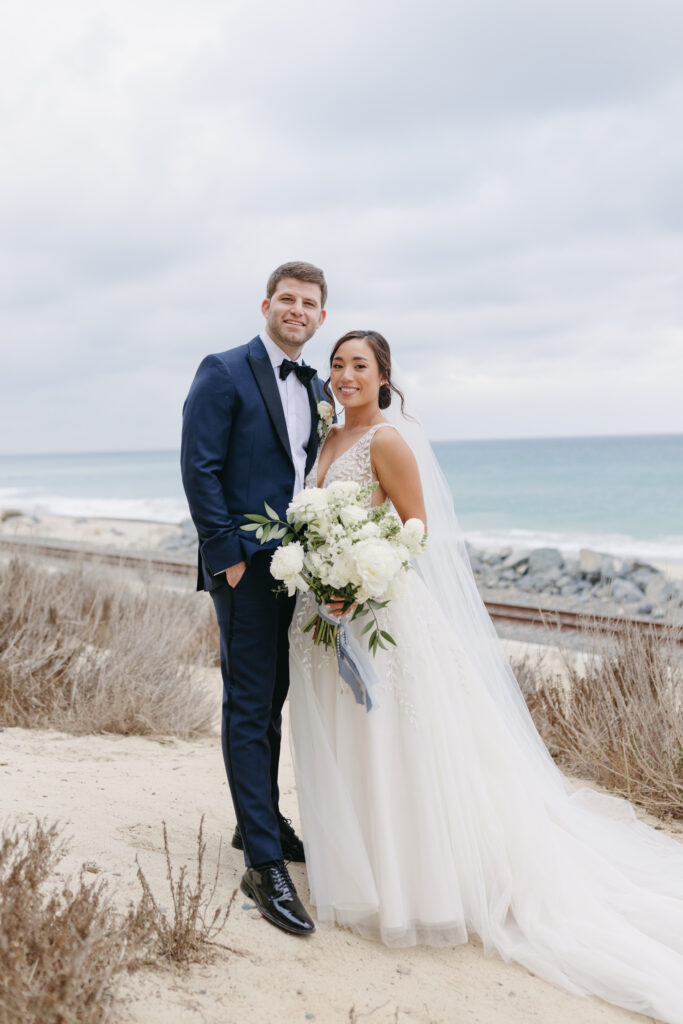 Groom wearing a navy blue suit with his bride holding a large bouquet with white flowers at the beach in San Clemente