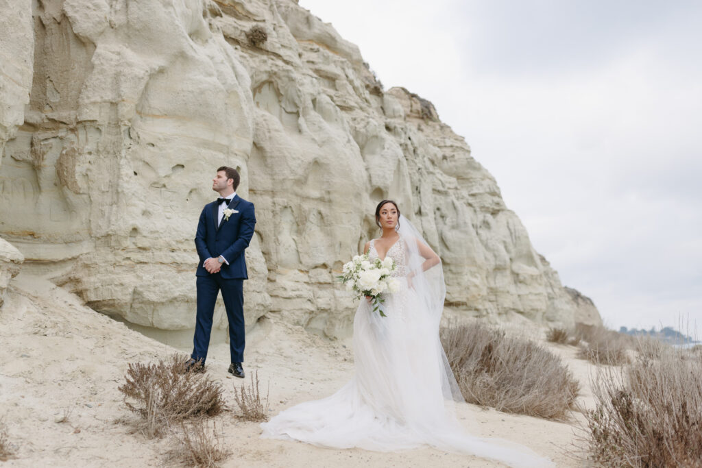 Bride with a long veil holding a large bouquet with white flowers standing next to her groom wearing a Navy blue suit at the beach in San Clemente