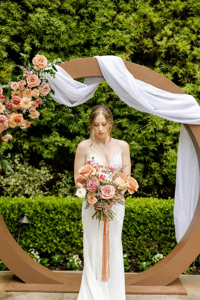 Bride holding a large bouquet with mauve flowers standing in front of a circle arch with mauve and peach flowers at Franciscan Gardens