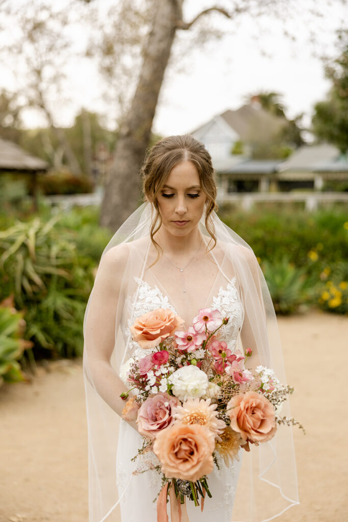 Bride with a long white veil looking down at her large bouquet with mauve pink and peach flowers in an outside garden in San Juan Capistrano