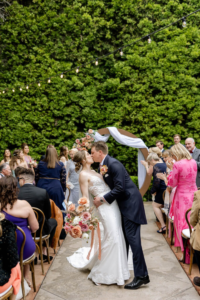 Groom wearing a navy blue suit kissing his bride wearing a lace white dress holding a large bouquet with mauve and peach flowers at Franciscan Gardens