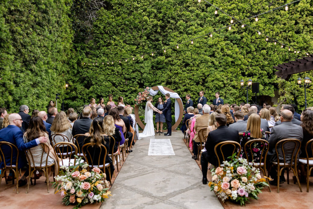Franciscan Gardens wedding ceremony couple standing at a circle arch draped with white fabric and accented with mauve and peach flowers with ground flower arrangements at the back of the aisle