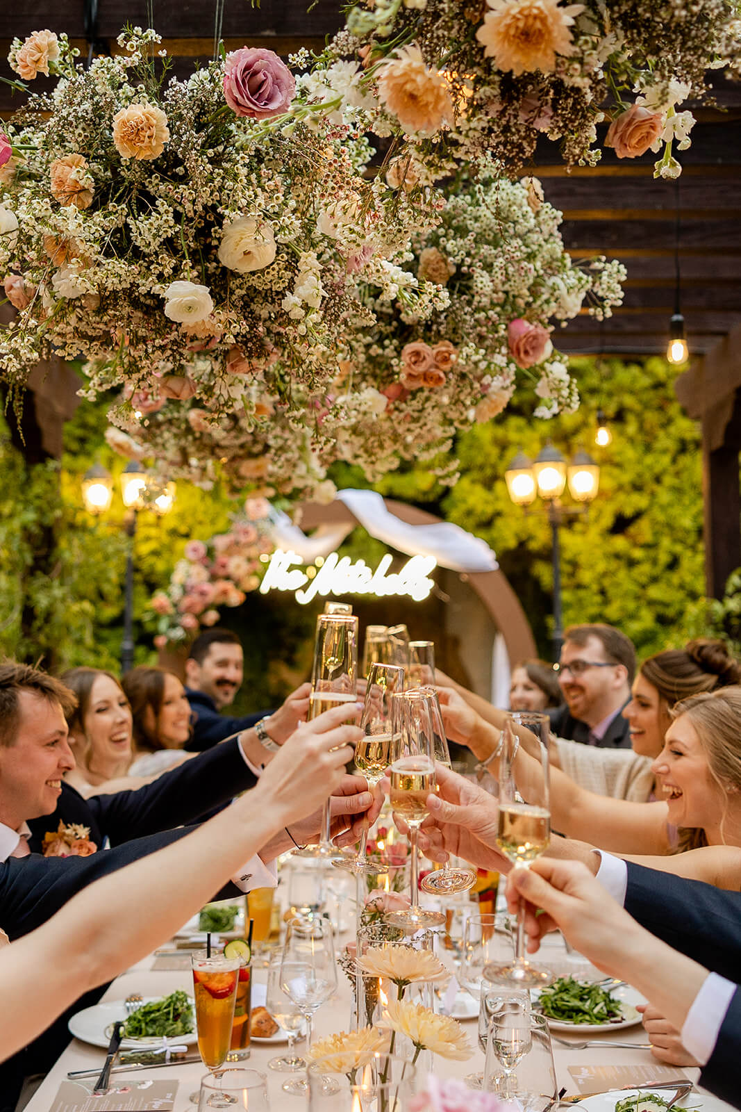 Wedding guests toast with champagne glasses at a long table under a hanging cloud floral installation at Franciscan Gardens