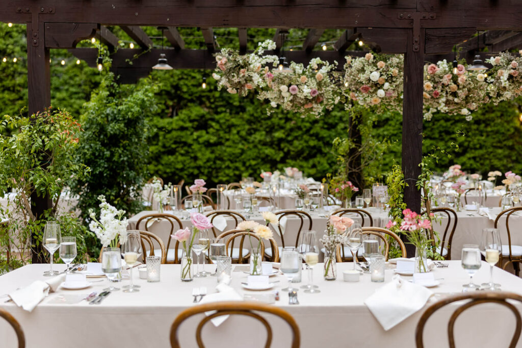 Rectangular wedding reception tables with mauve and peach bud vase centerpieces in front of a dark wood pergola with a hanging floral cloud installation at Franciscan Gardens