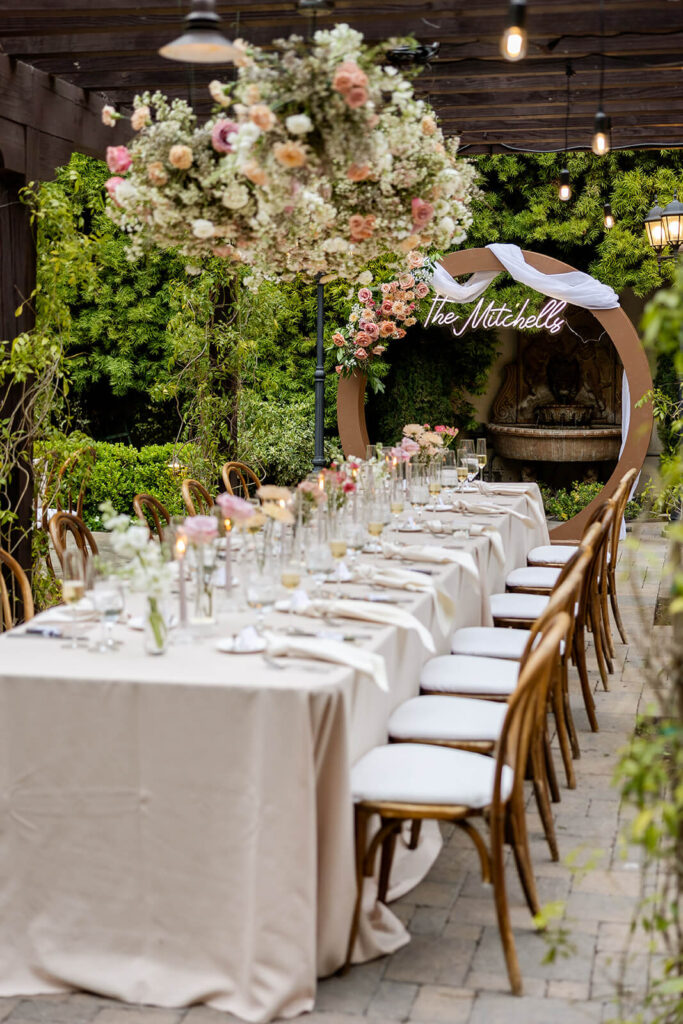 Long rectangular head table under a dark wood pergola with hanging cloud floral installation above the table with bud vase centerpieces at Franciscan Gardens