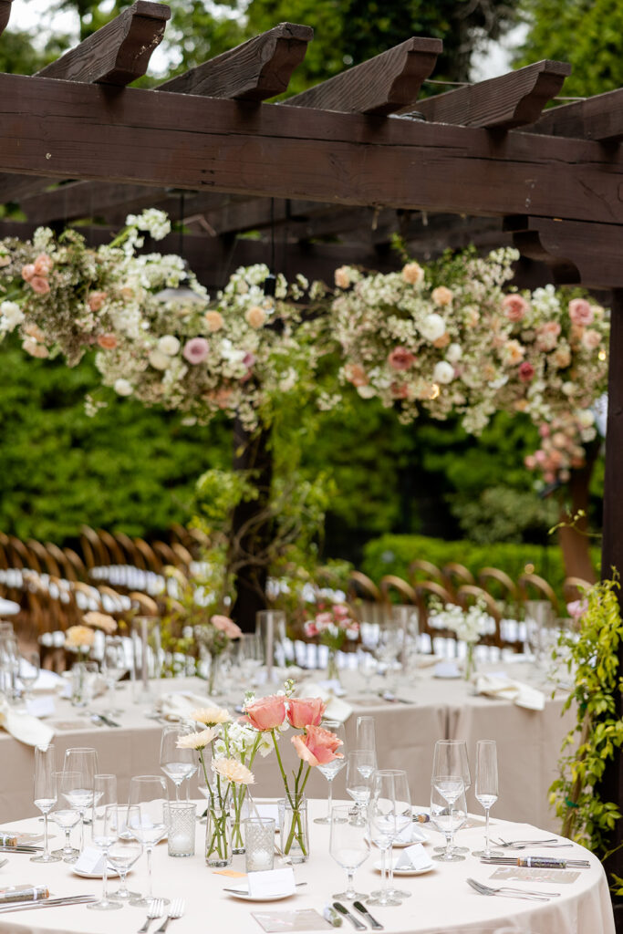 Hanging floral cloud installation on a dark wood pergola with a round wedding reception table with bud vase centerpieces
