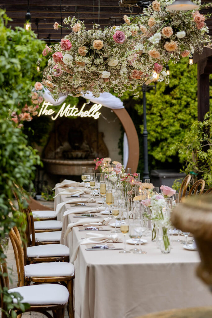 Hanging floral cloud installation over a long rectangular table with bud vases centerpieces and a circle arch at the end of the table with a neon sign reading The Mitchells at Franciscan Gardens