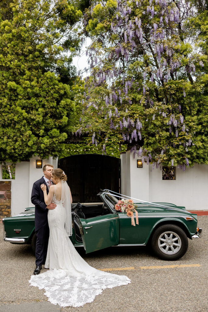 Newlywed couple standing outside Franciscan Gardens with a dark green classic convertible car topped with a mauve and peach bouquet