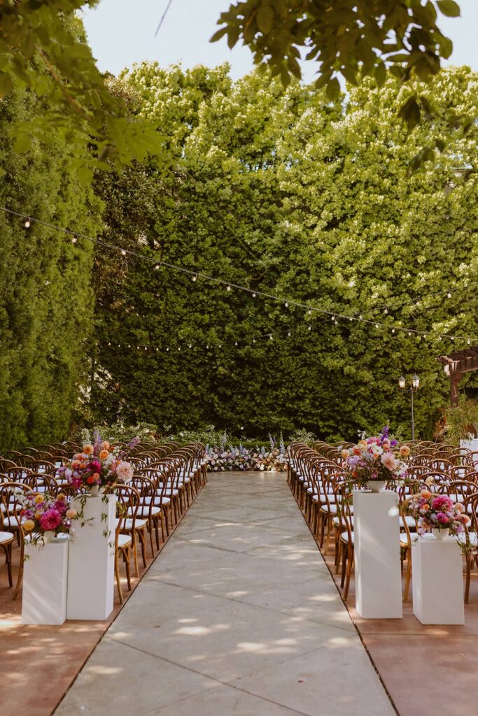 White pillars at the aisle with colorful floral centerpieces with a growing garden meadow at the end of the aisle