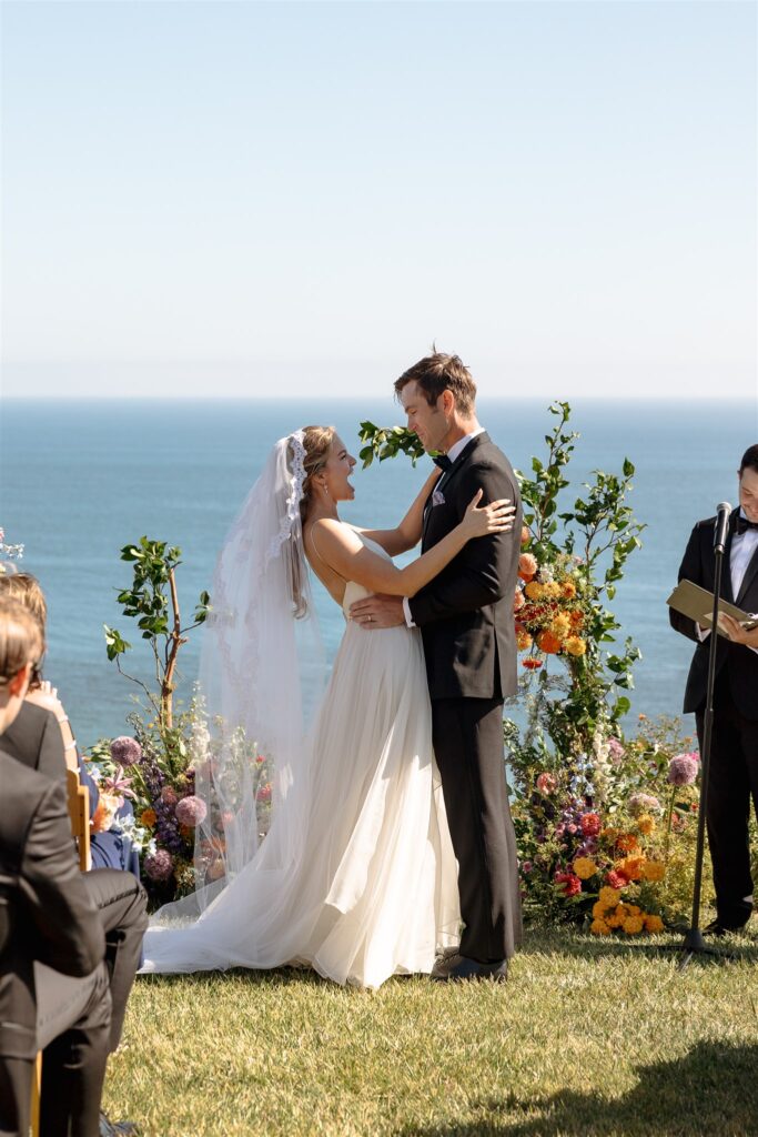 bride and groom in front of wedding ceremony flower installation with ocean in background